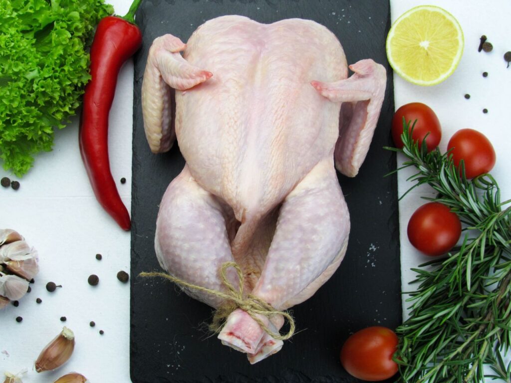 Overhead shot of a raw whole chicken surrounded by fresh vegetables and spices on a slate board.